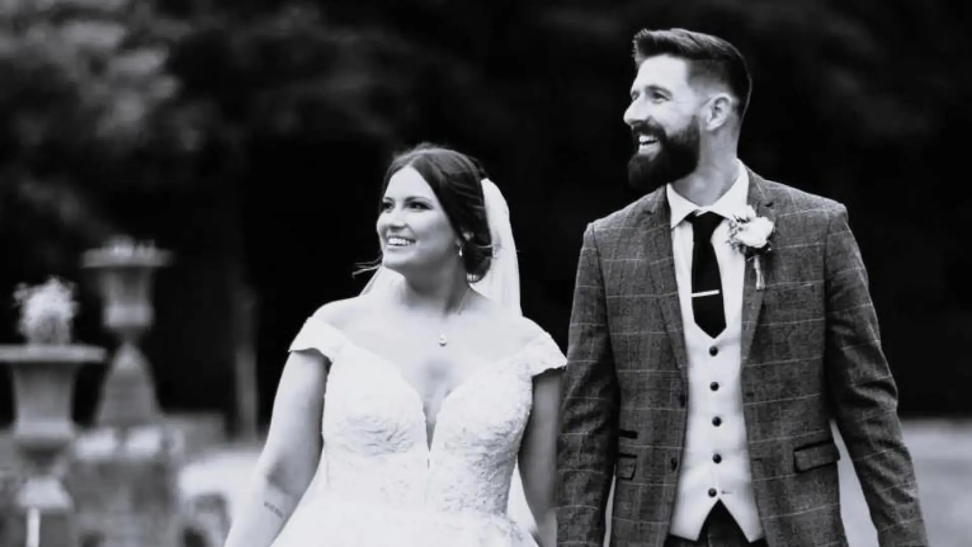 Joshua Vickers (27) im Anzug und seine Frau Laura in einem Hochzeitskleid gehen über einen Weg in einem Park.