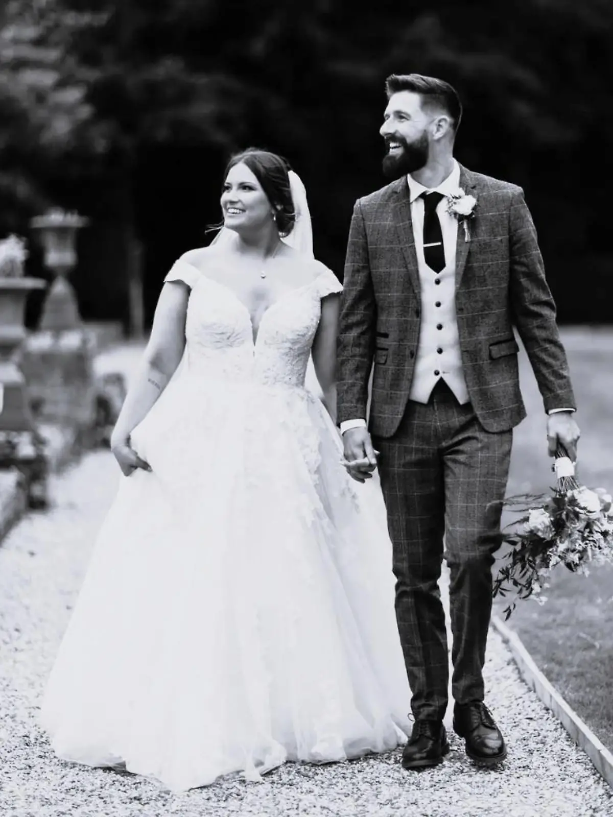Joshua Vickers (27) im Anzug und seine Frau Laura in einem Hochzeitskleid gehen über einen Weg in einem Park.