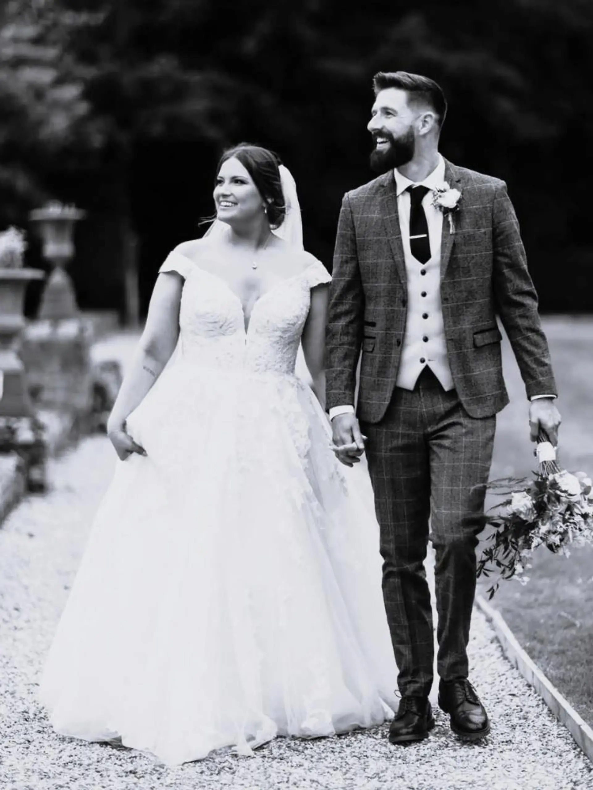 Joshua Vickers (27) im Anzug und seine Frau Laura in einem Hochzeitskleid gehen über einen Weg in einem Park.