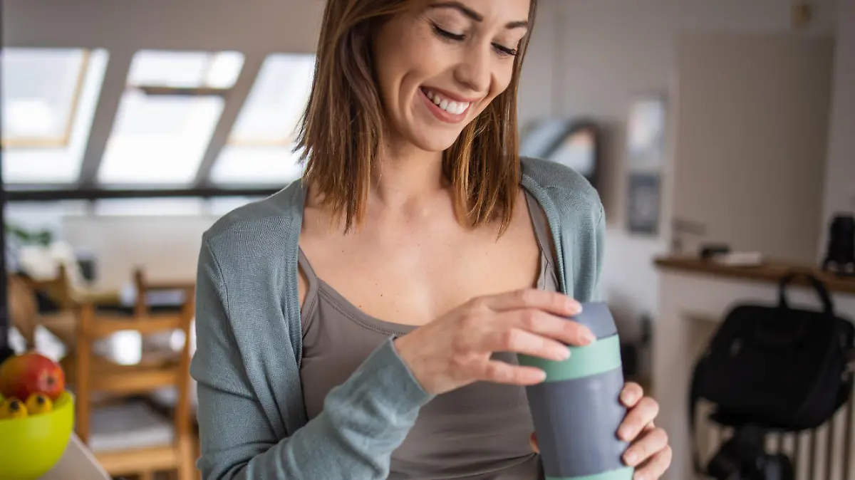Beautiful smiling woman standing in kitchen and holding reusable travel mug in hands