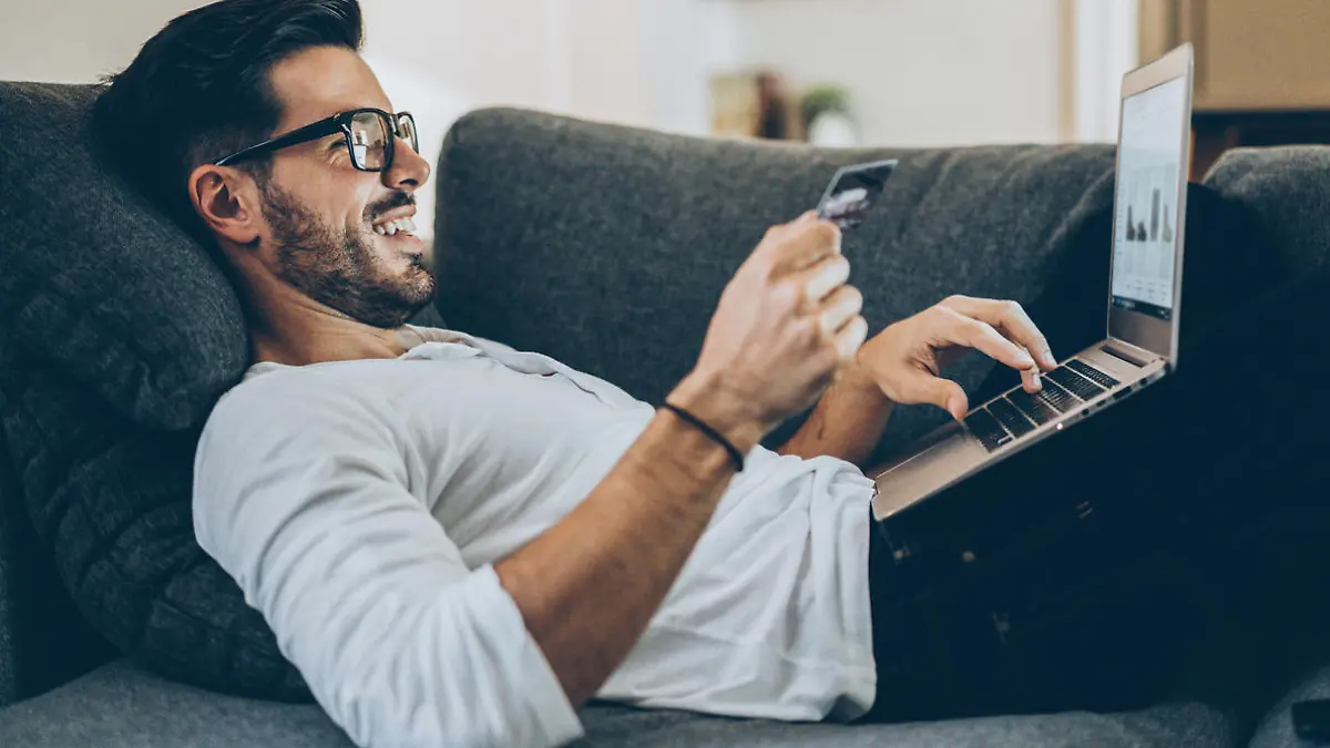 Smiling man lying on the couch and shopping online with credit card and laptop