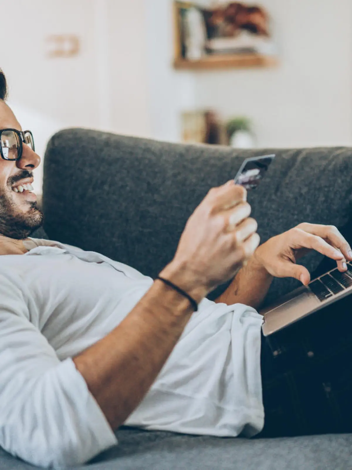 Smiling man lying on the couch and shopping online with credit card and laptop