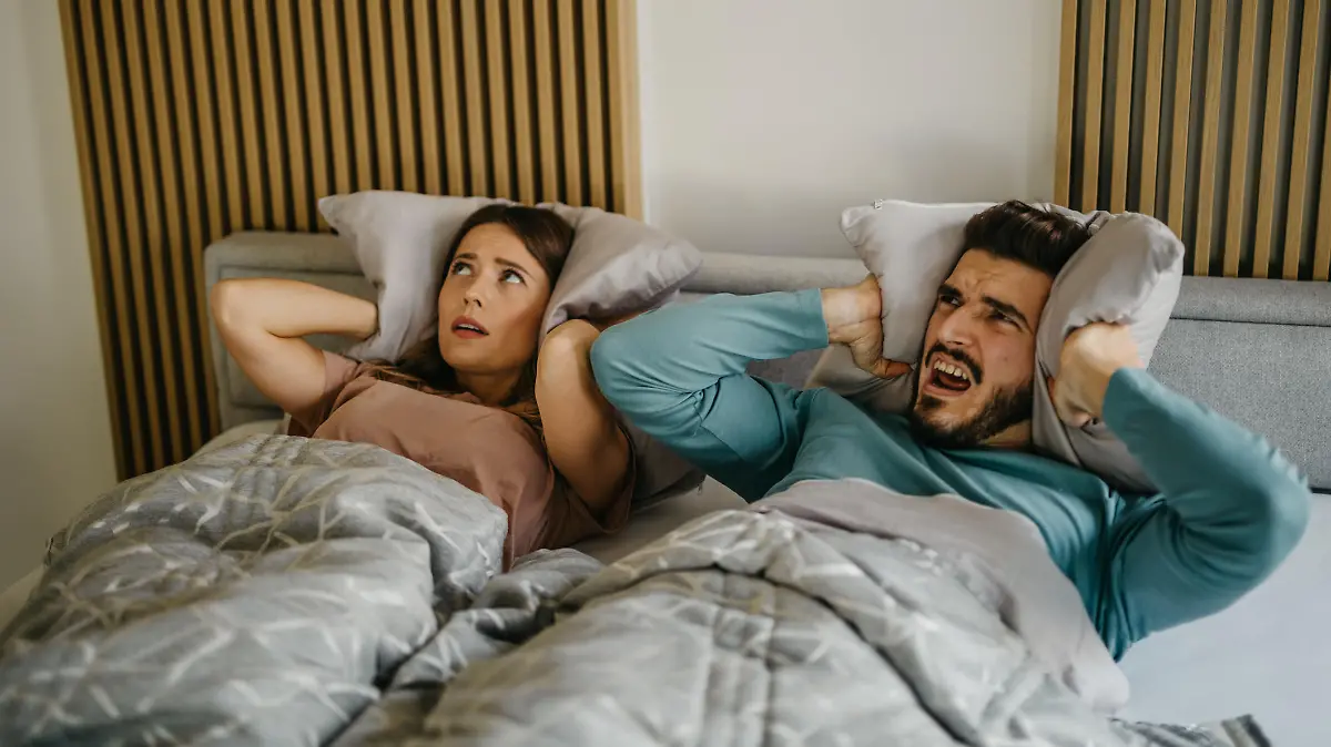 Annoyed adult woman and man in bed suffering neighbor noise at home. The couple has an unhappy facial expression closing their ears with pillows while lying in bed.