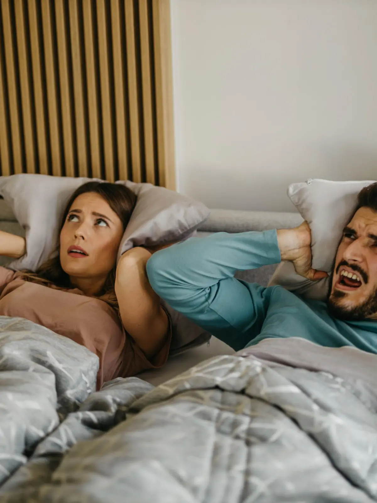Annoyed adult woman and man in bed suffering neighbor noise at home. The couple has an unhappy facial expression closing their ears with pillows while lying in bed.