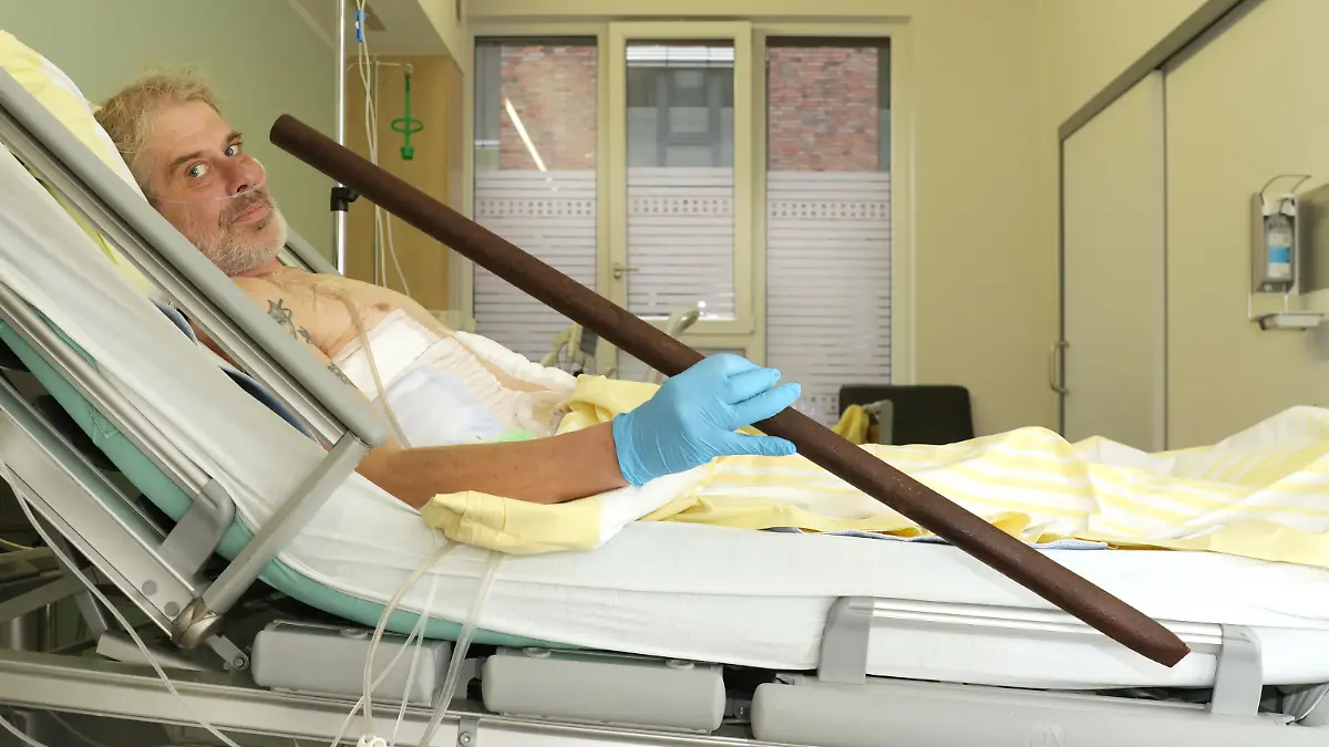 Ein Mann liegt in einem Krankenhausbett, er hält eine Stange in der Hand.