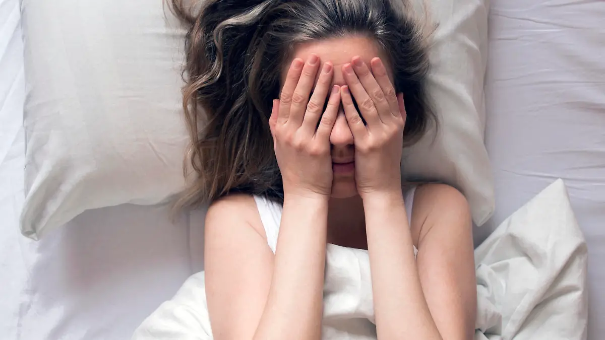 Girl long haired brunette lying on back on white bed having insomnia and covering hide face hands. Early awakened. Naughty sunny morning. top view, feeling sad or depressed