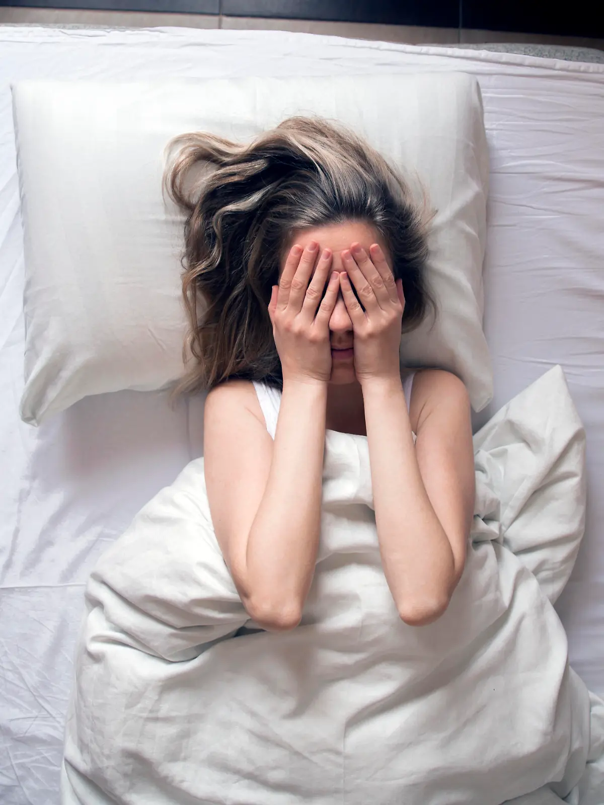 Girl long haired brunette lying on back on white bed having insomnia and covering hide face hands. Early awakened. Naughty sunny morning. top view, feeling sad or depressed
