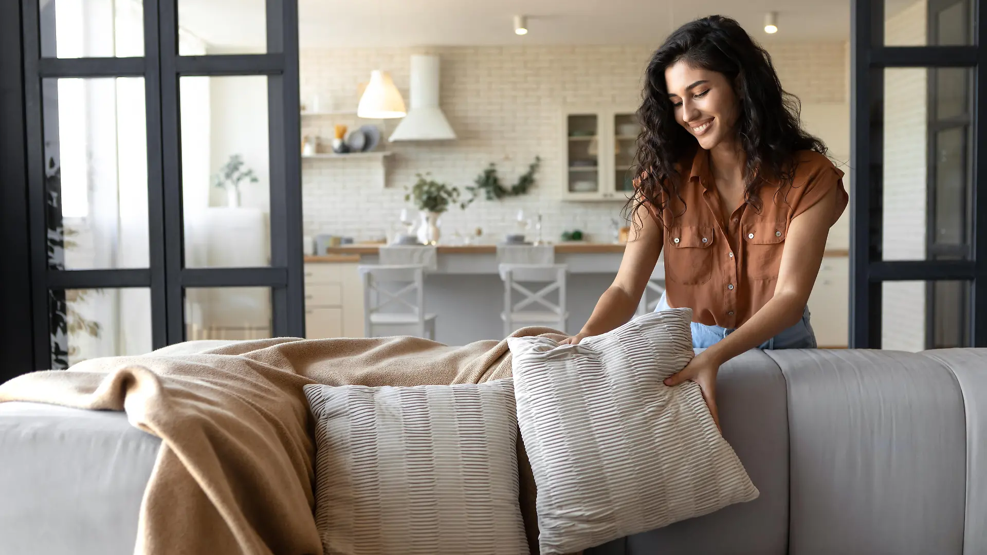 Lovely young woman putting soft pillows and plaid on comfy sofa, making her home cozy and warm, copy space. Millennial lady decorating her couch with cushions and blanket for autumn or winter