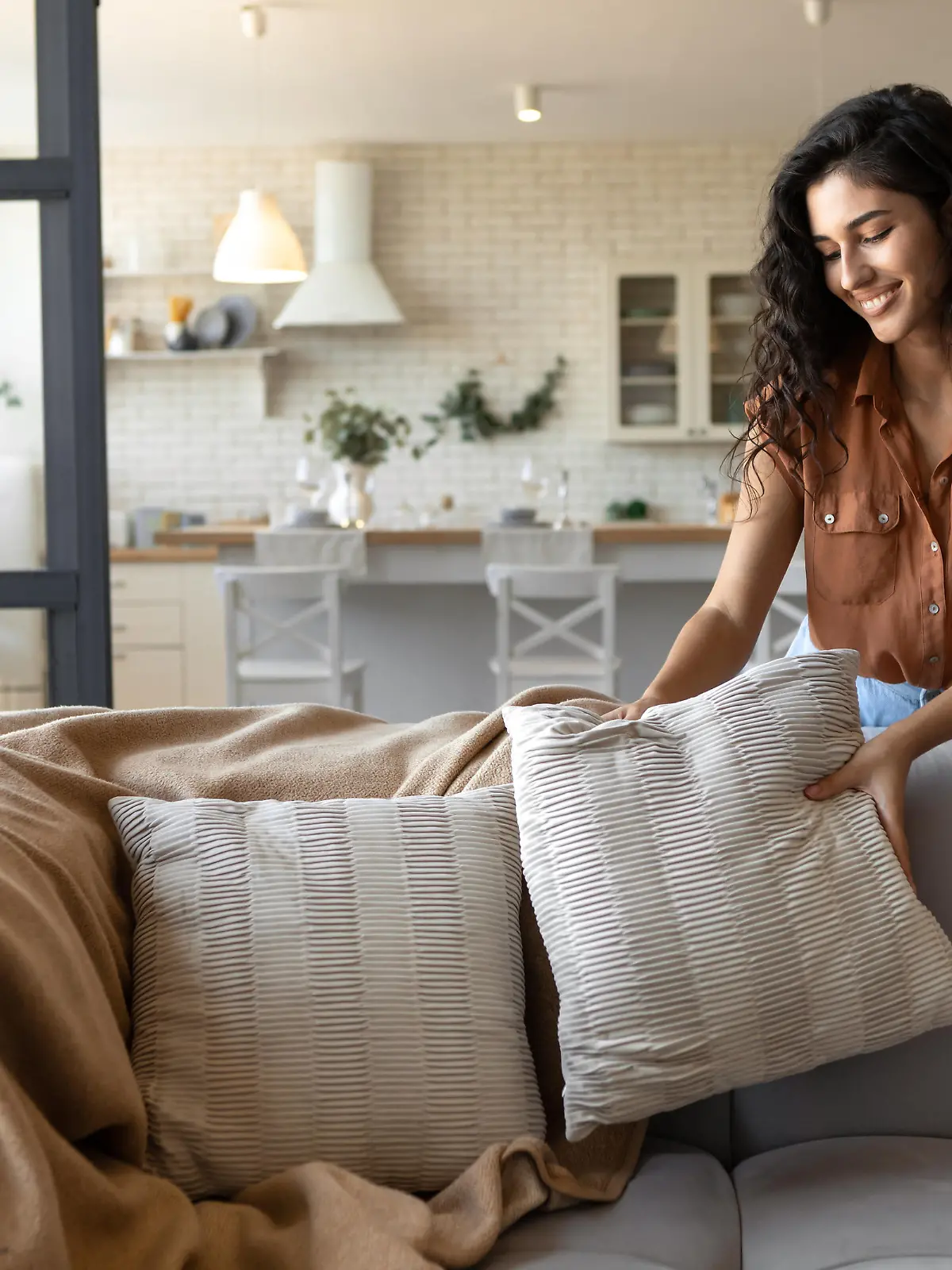 Lovely young woman putting soft pillows and plaid on comfy sofa, making her home cozy and warm, copy space. Millennial lady decorating her couch with cushions and blanket for autumn or winter