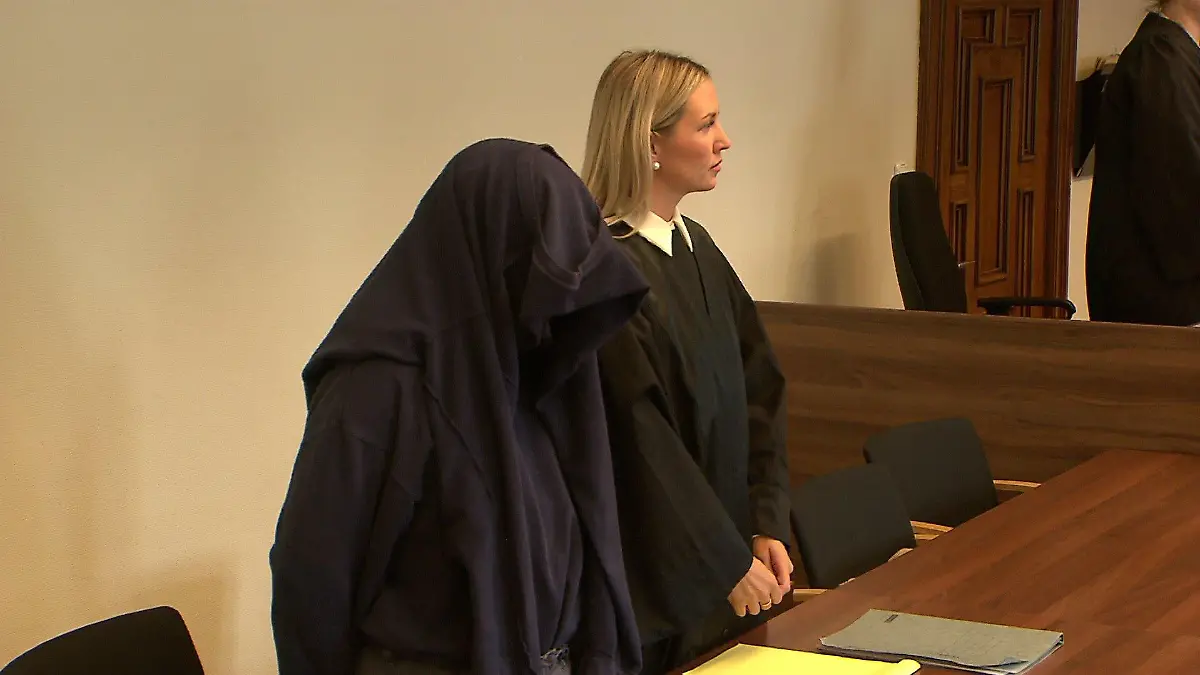 Ein mit einem Pullover vermummter Mann steht an einer Anklagebank.