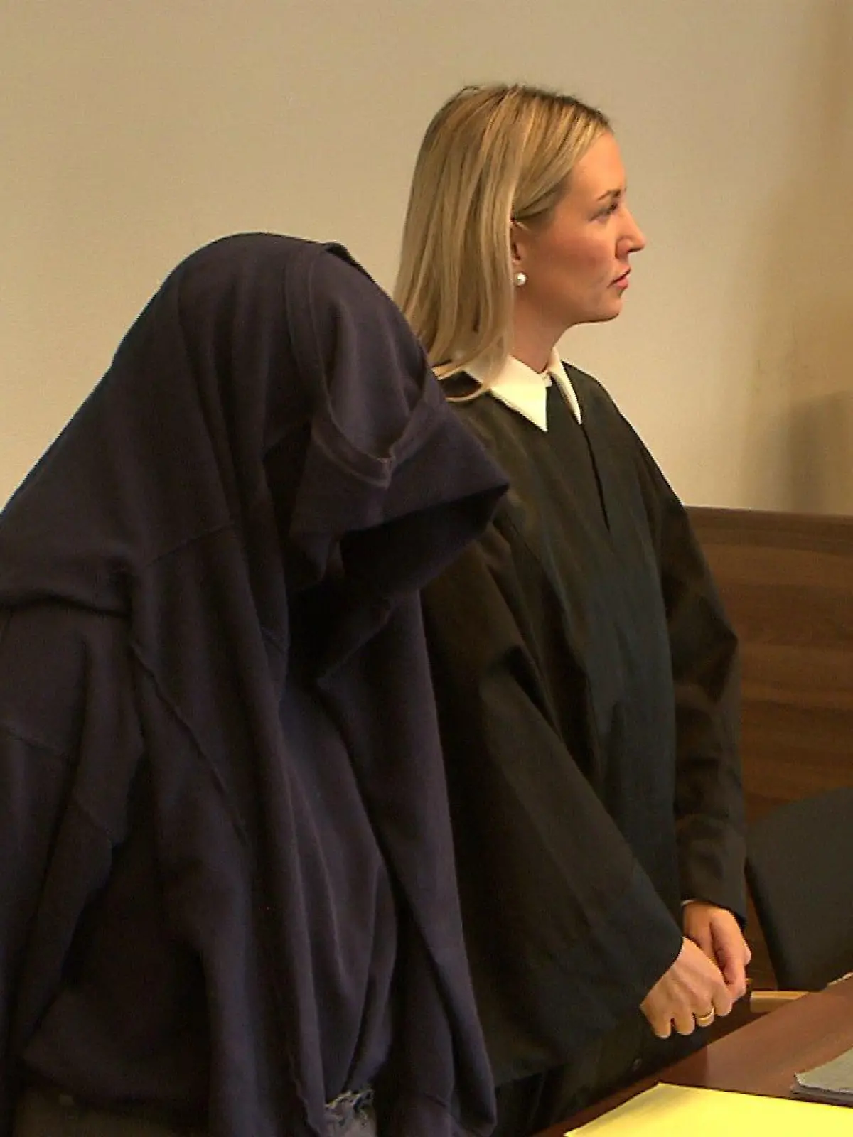 Ein mit einem Pullover vermummter Mann steht an einer Anklagebank.
