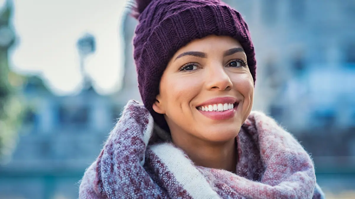 Closeup face of a young happy african woman enjoying winter wearing scarf and cap. Smiling brazilian girl in a colorful shawl looking at camera in a cold day. Multiethnic woman with knitted bordeaux hat and woolen scarf.