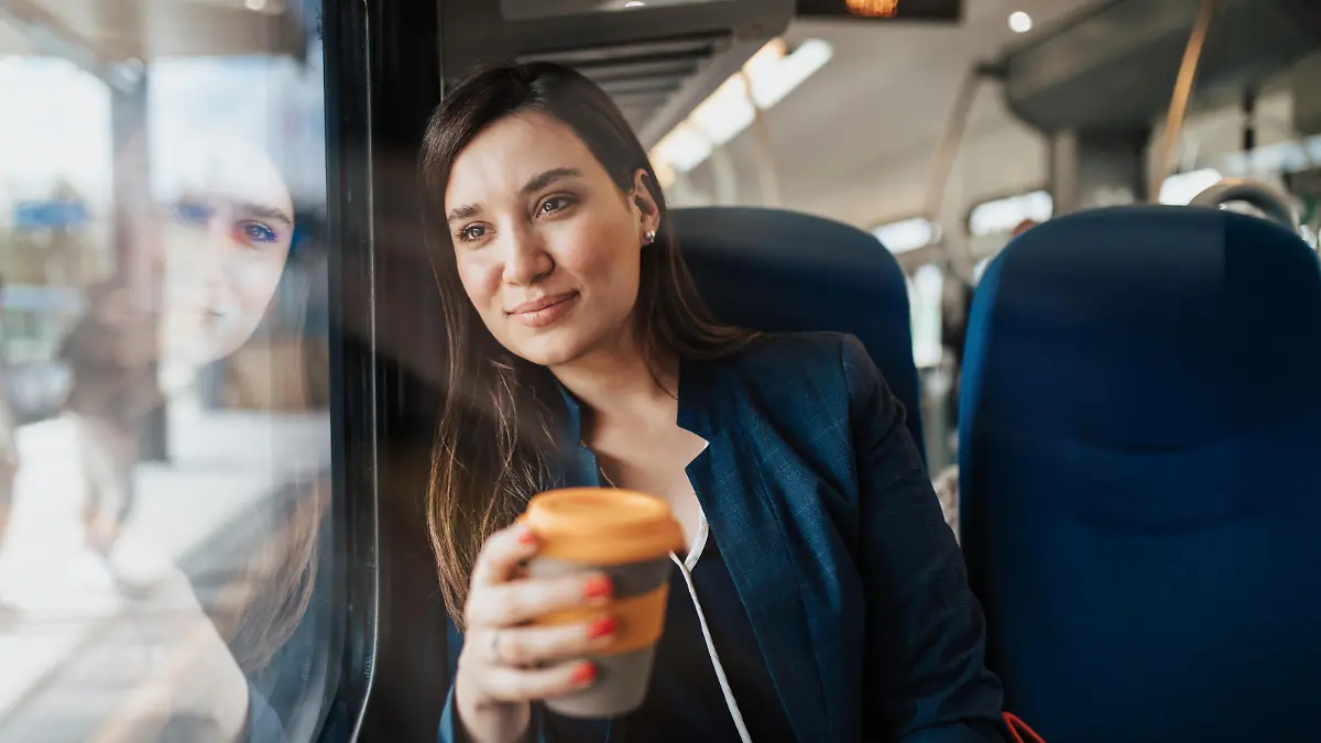 A beautiful young business woman is sitting in the cab of a high-speed train, looking through window and drinking coffee to go.