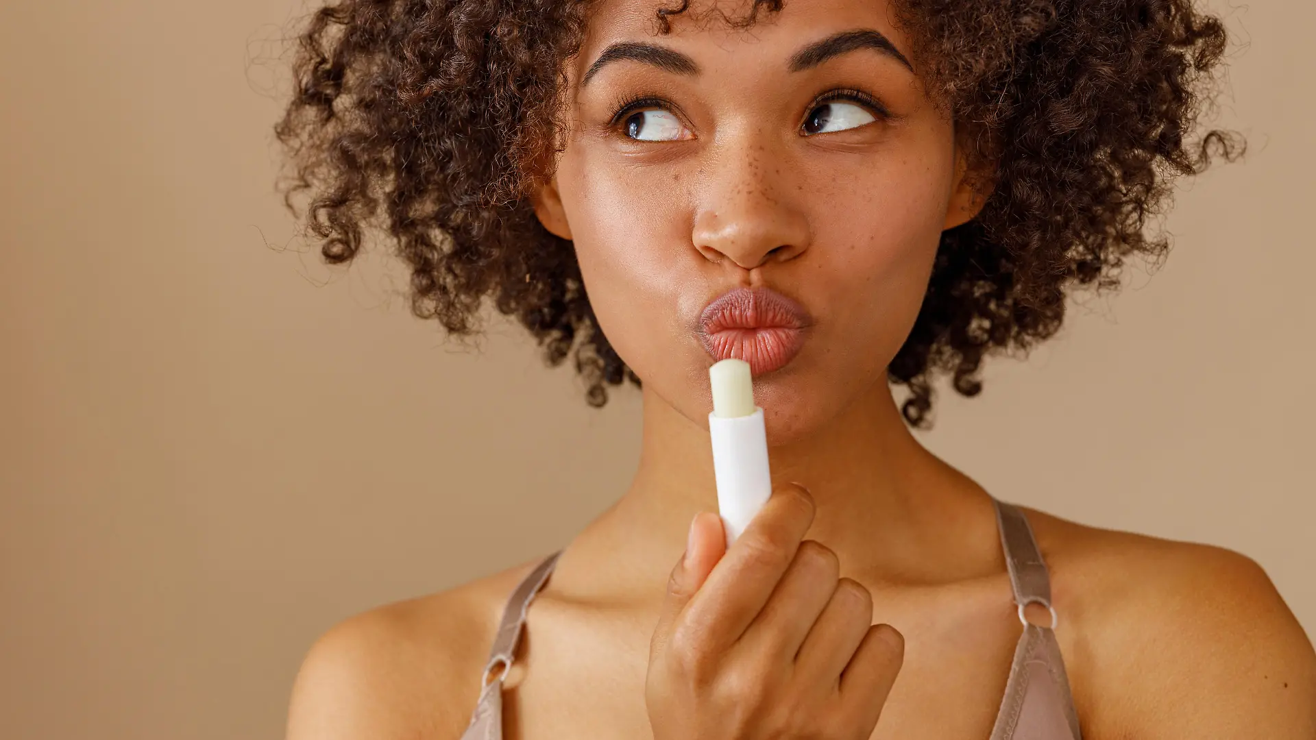 Beautiful young model holding cosmetic lip balm and flirting, looking sideways with teasing expression of lips on pastel beige background