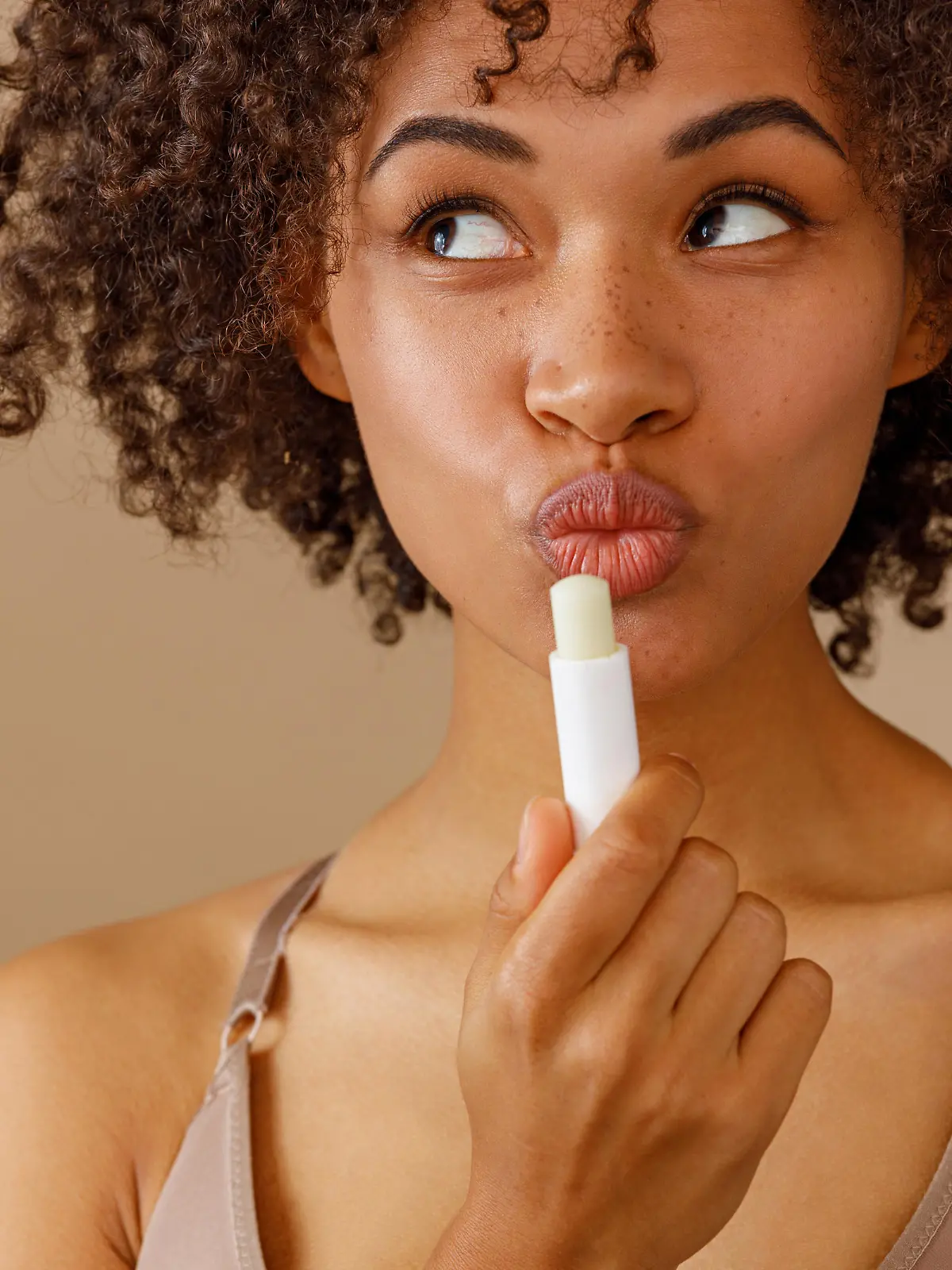 Beautiful young model holding cosmetic lip balm and flirting, looking sideways with teasing expression of lips on pastel beige background