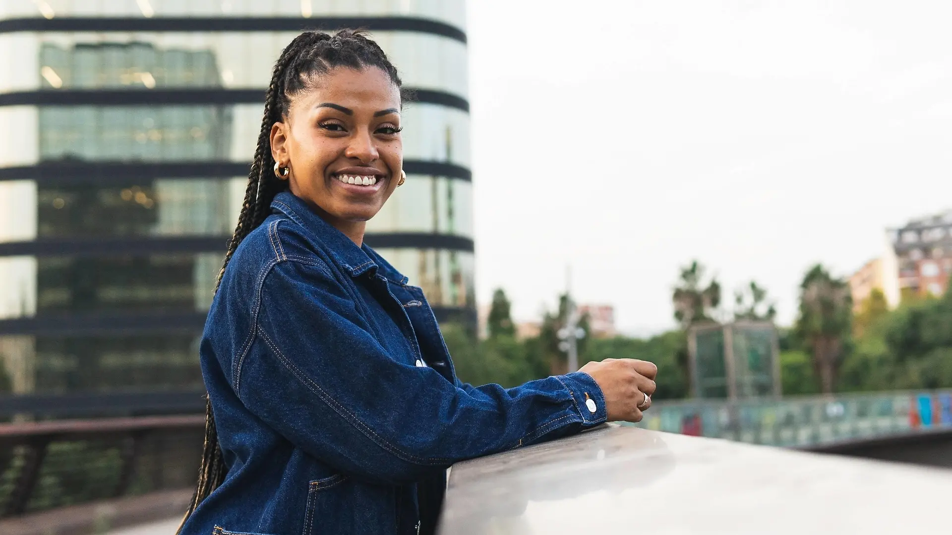 Low angle of positive African American female in denim jacket smiling and looking at camera while leaning on railing standing on bridge against cityscape DavidMunoz_ID4111_134841_049 Copyright: xDavidxMunozx