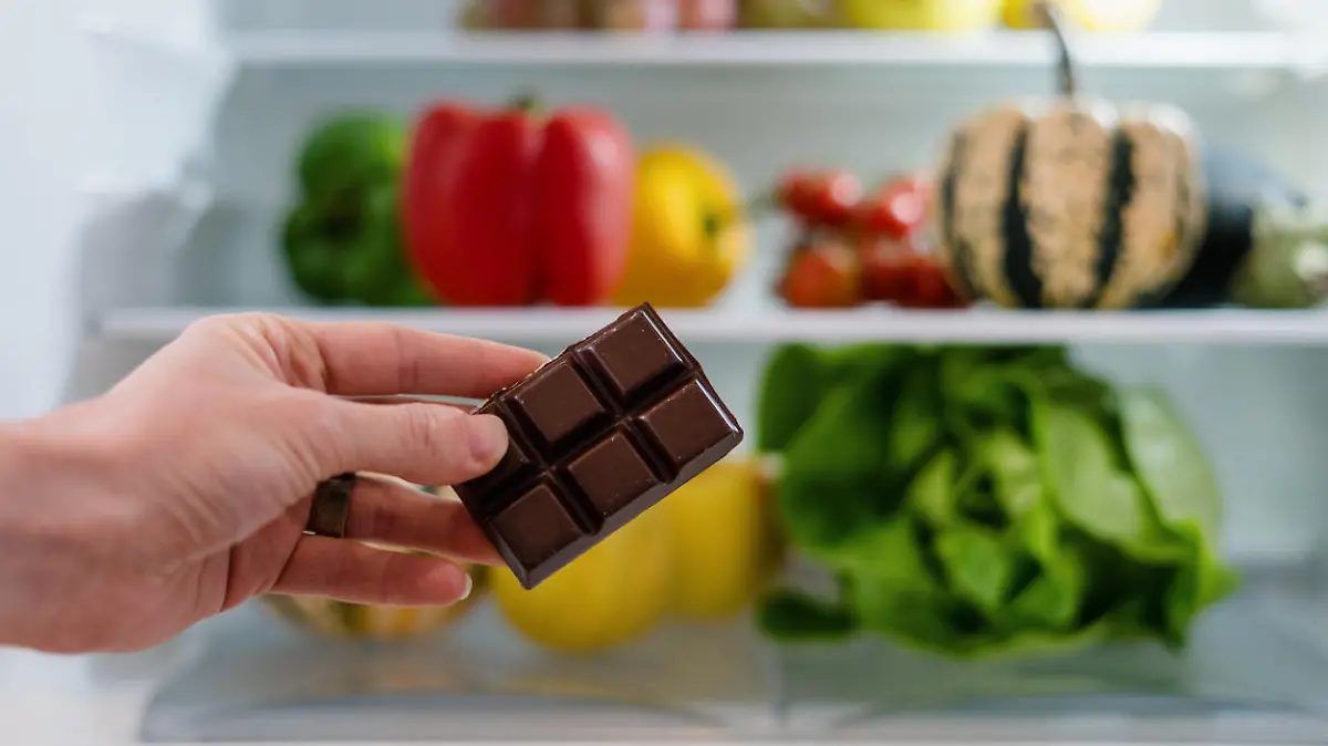 fresh fruits and vegetables on shelf in open fridge and female hand with sweets, healthy eating concept, choice between healthy and unhealthy food