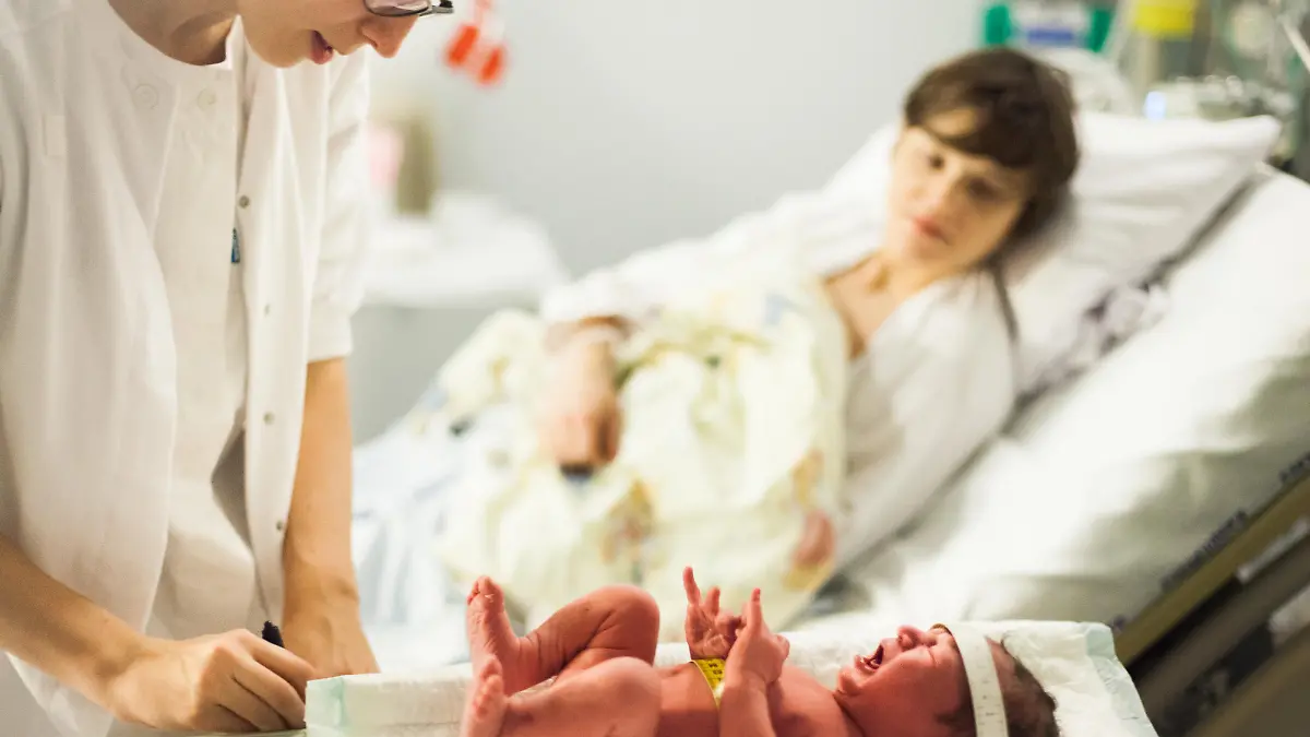 Calm and patient midwife weighs a screaming and crying newborn baby, mother in background