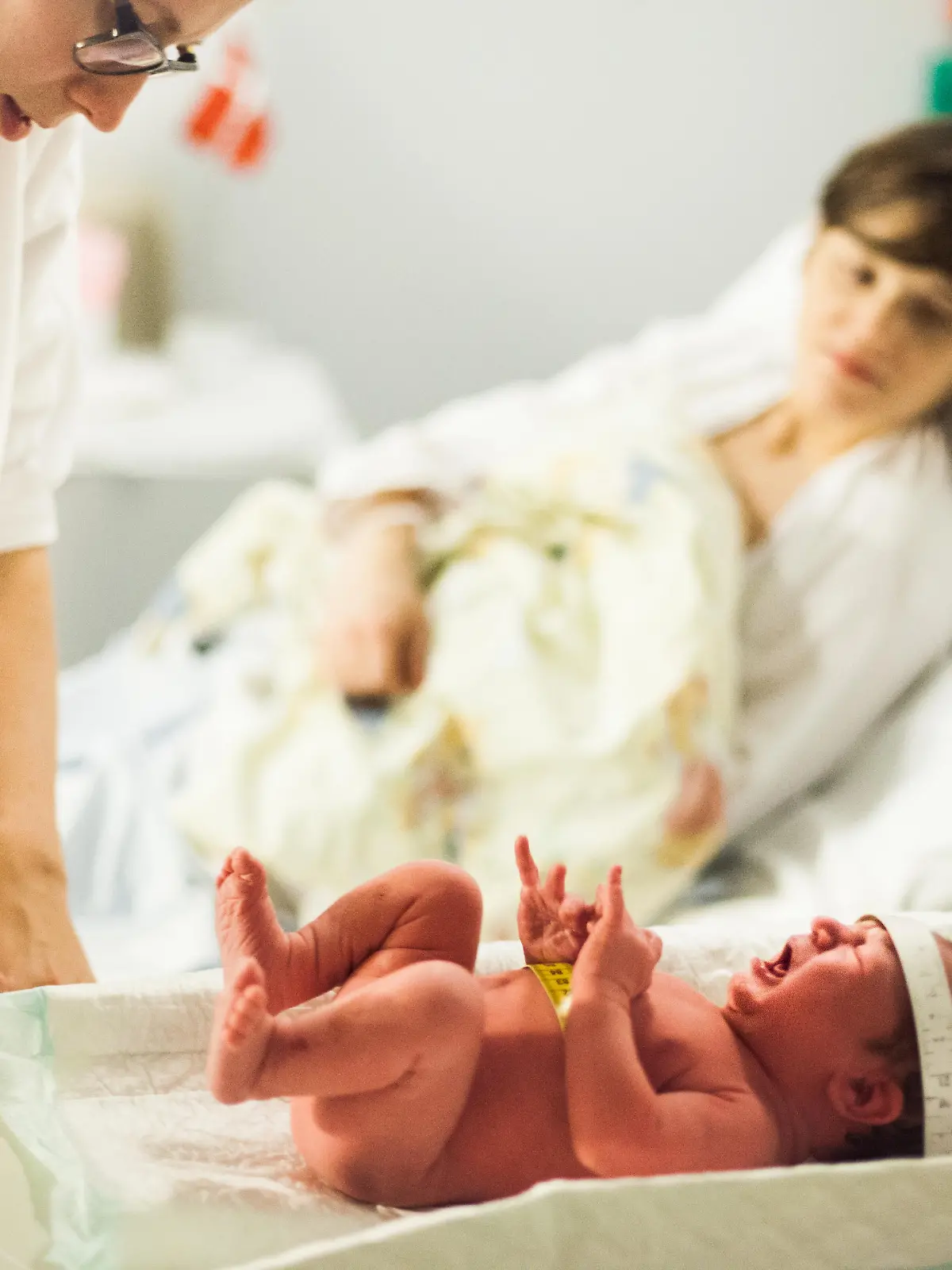 Calm and patient midwife weighs a screaming and crying newborn baby, mother in background