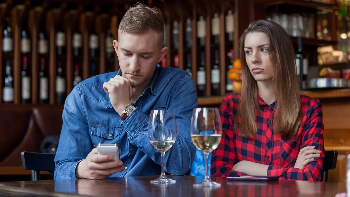 Sulking woman sitting next to man reading text messages in restaurant