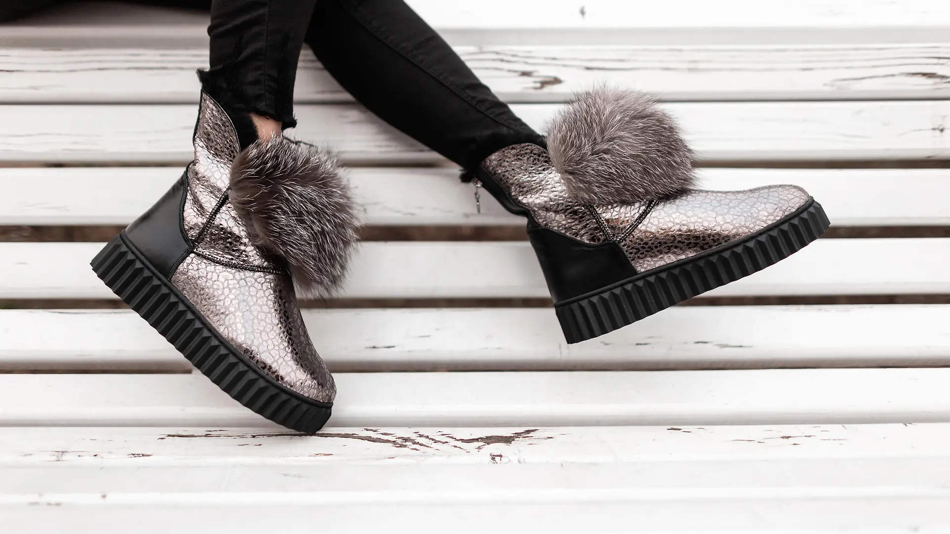 Beautiful silver shiny winter shoes with gray fur on female legs. New collection of fashionable warm glamorous boots. Modern design. Seasonal fashion. Close-up.