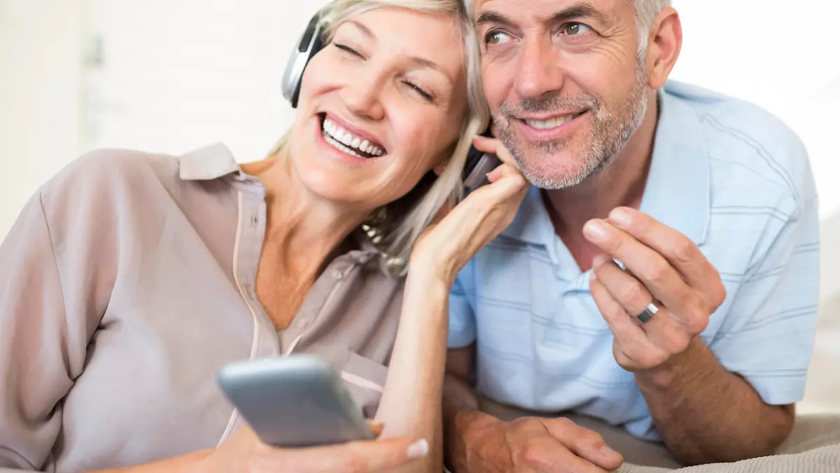 Cheerful mature man and woman with headphones and cellphone sitting on sofa at home