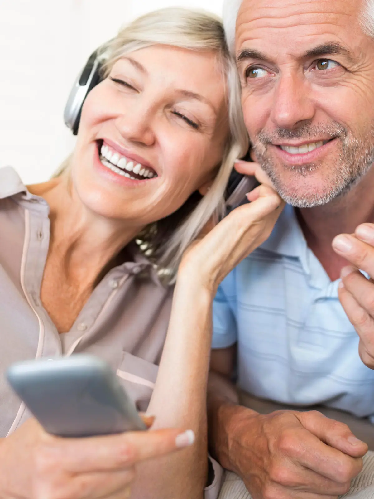Cheerful mature man and woman with headphones and cellphone sitting on sofa at home