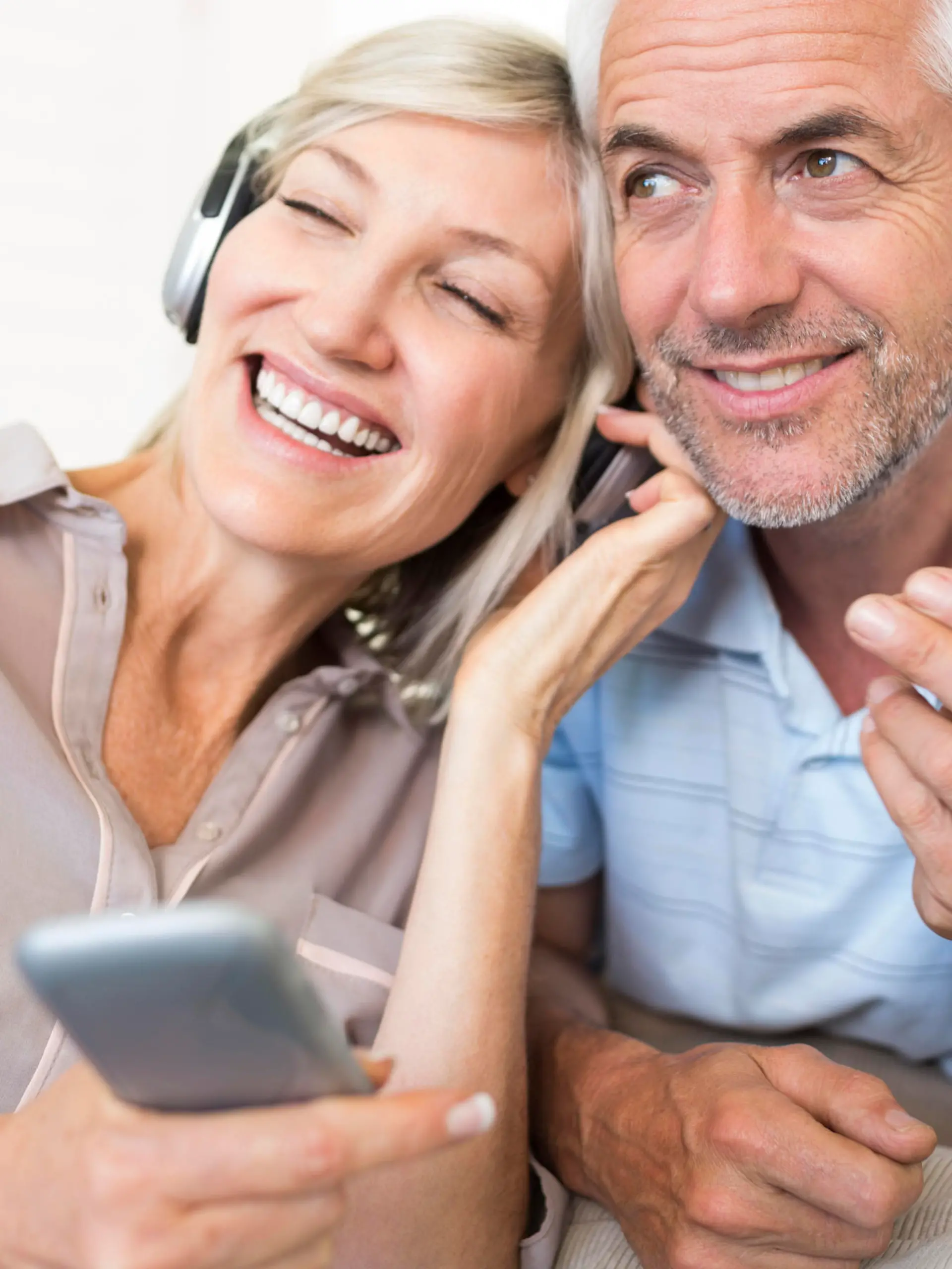 Cheerful mature man and woman with headphones and cellphone sitting on sofa at home