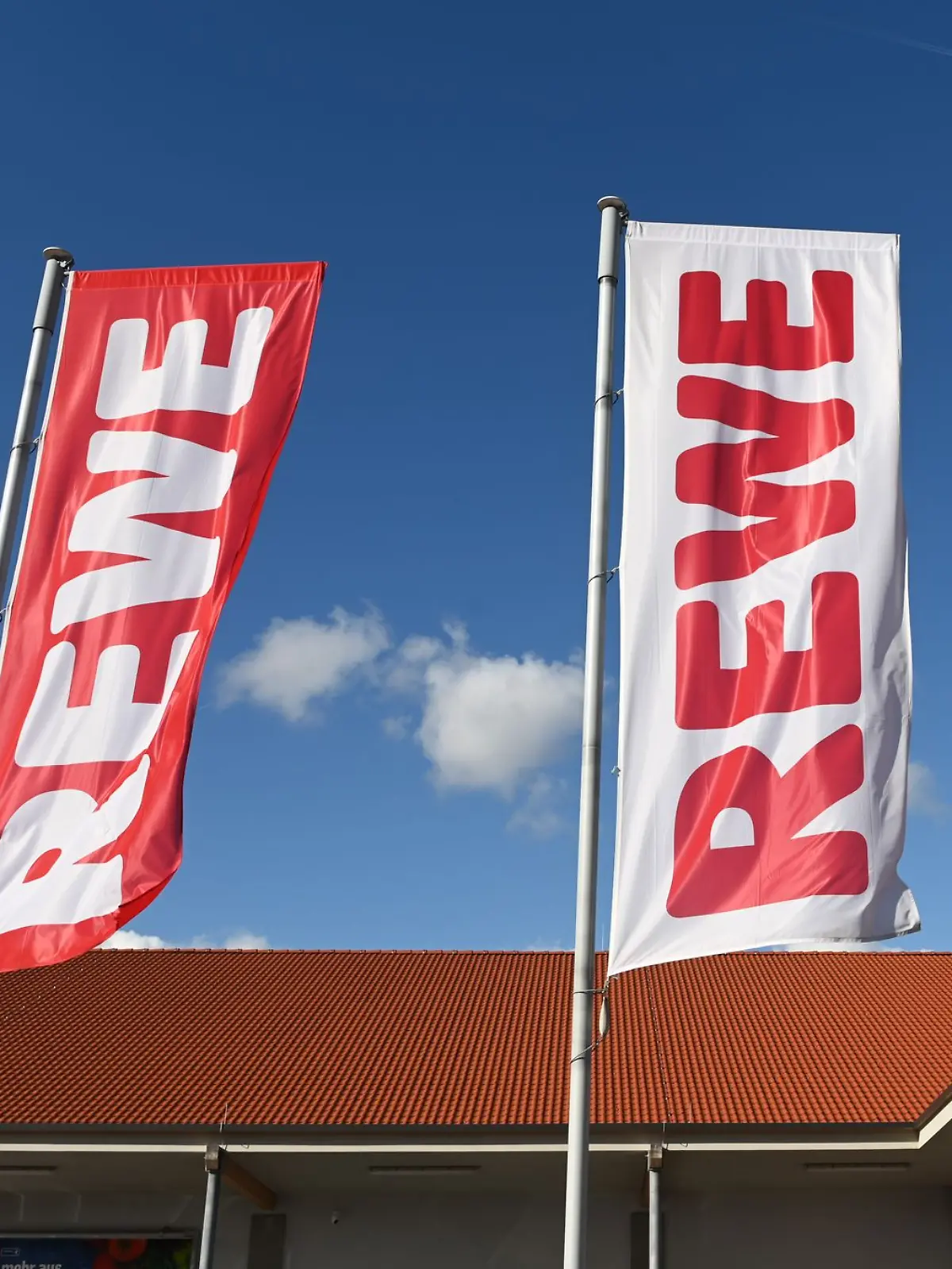Fahnen mit dem Logo des Lebensmitteleinzelhändlers Rewe wehen vor einer Filiale.