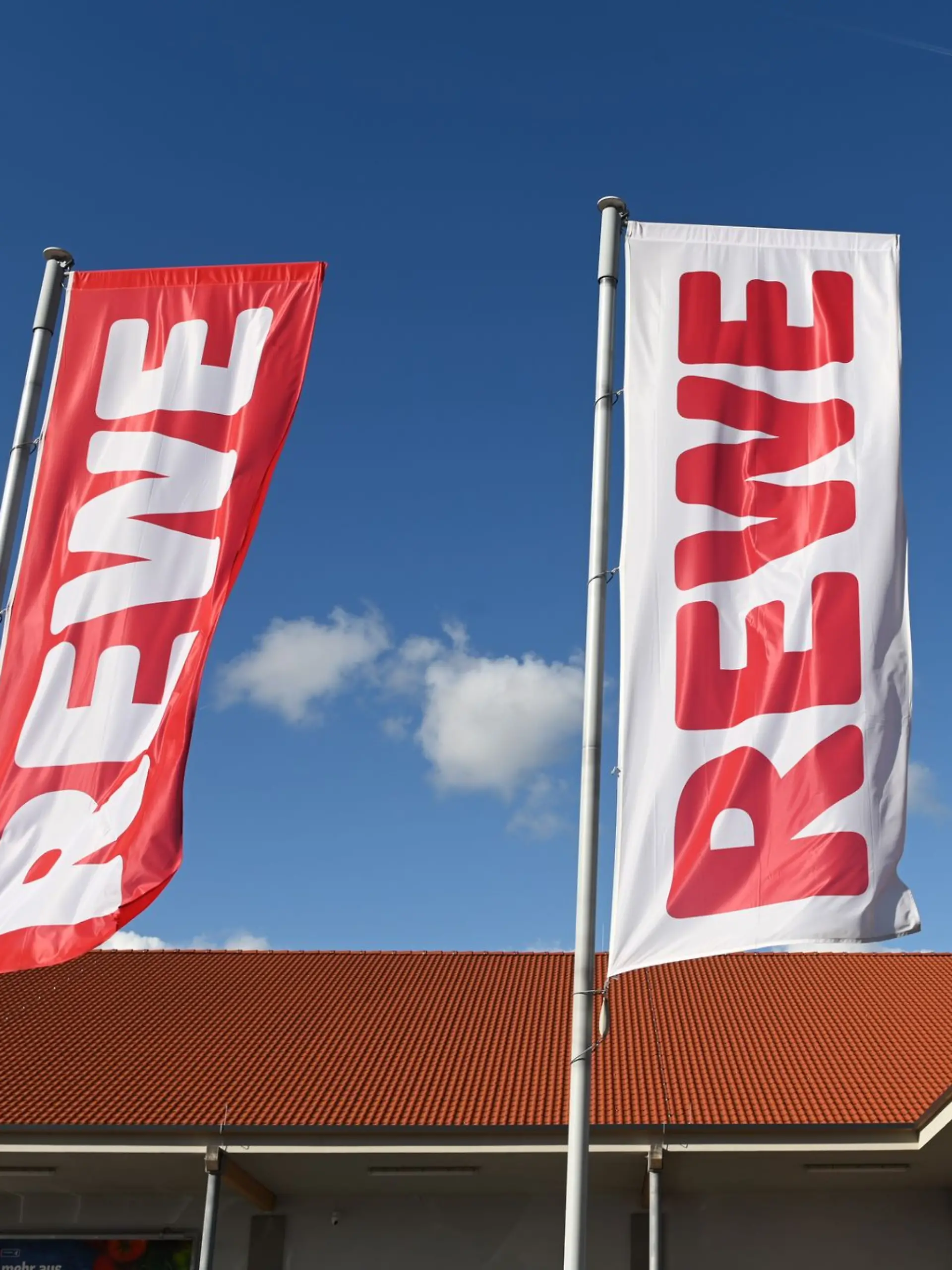 Fahnen mit dem Logo des Lebensmitteleinzelhändlers Rewe wehen vor einer Filiale.