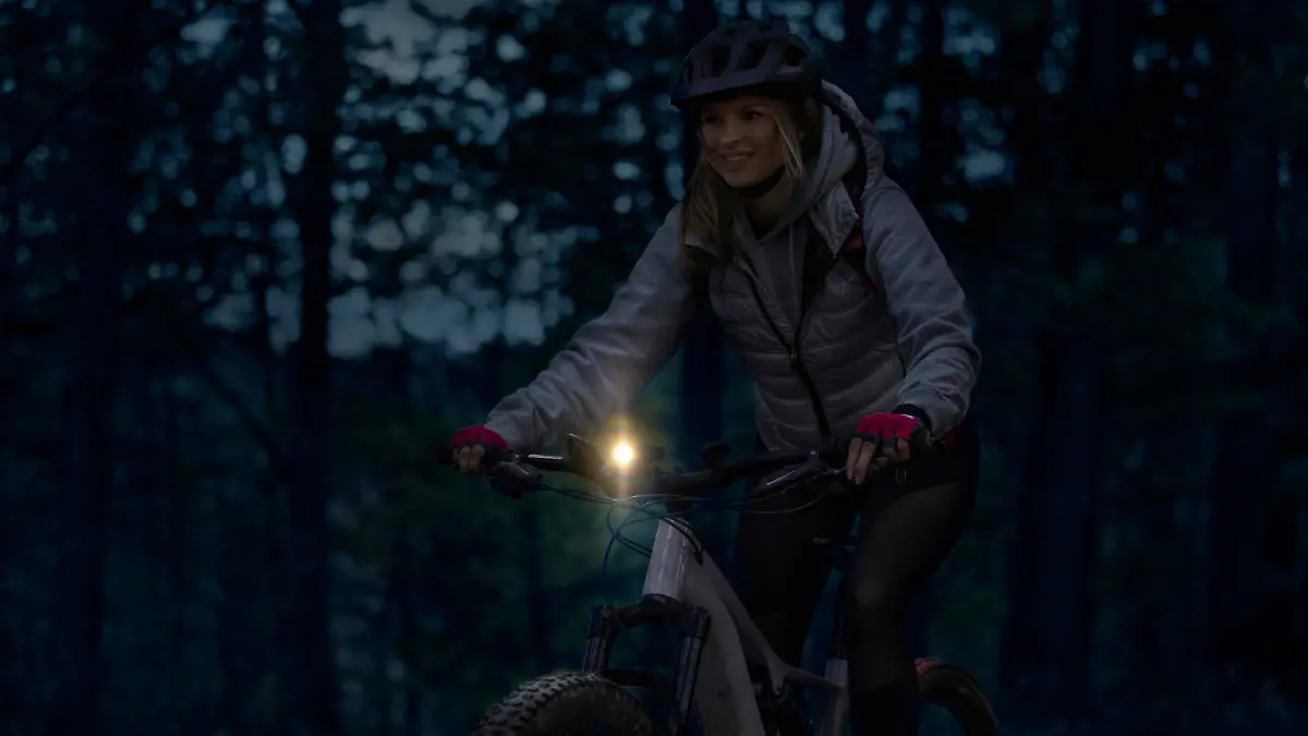 Low-key photo of a cheerful young woman riding her enduro mountain bike (eMTB) at night, with a front light on.