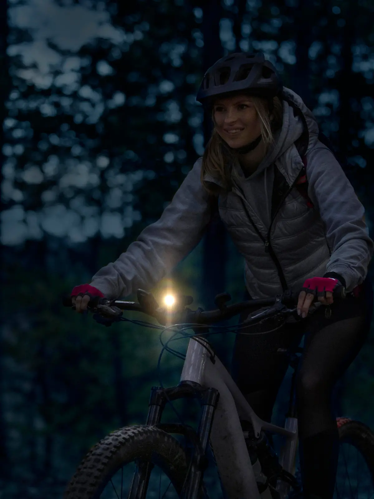 Low-key photo of a cheerful young woman riding her enduro mountain bike (eMTB) at night, with a front light on.