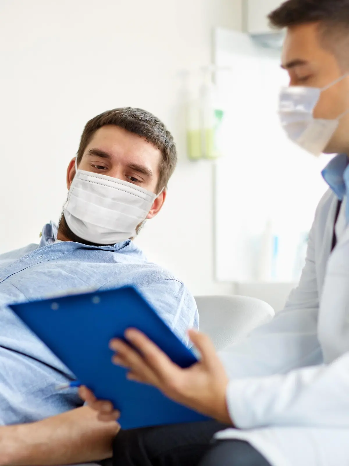 medicine, healthcare and pandemic concept - doctor wearing face protective medical mask for protection from virus disease with clipboard and young male patient at medical exam at hospital