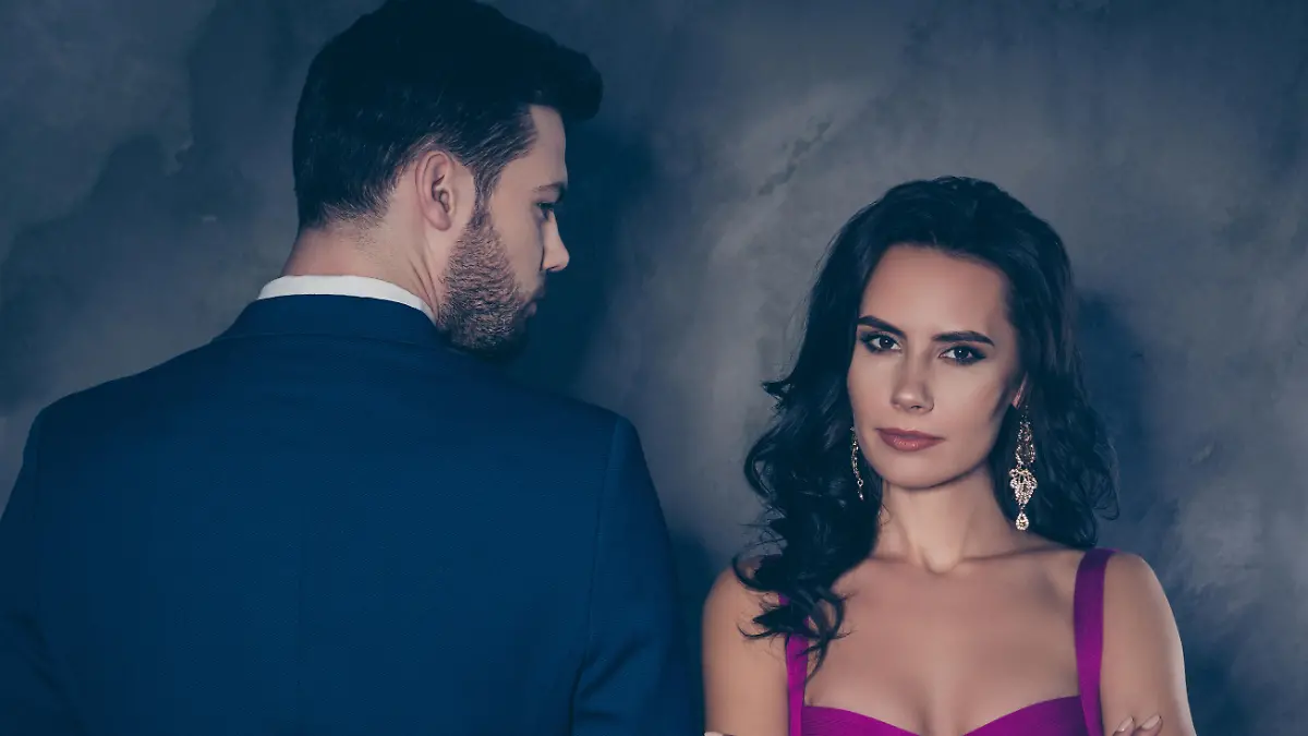 Rear view portrait of brunet business gentleman looking at serious lady with front view in purple outfit with jewelry curly hair seductive decollete holding arms crossed isolated on grey background