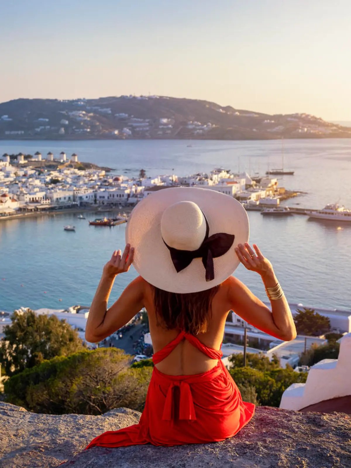 A elegant tourist woman in a red dress enjoys the view over the city of Mykonos island during a golden summer sunset, Cyclades, Greece