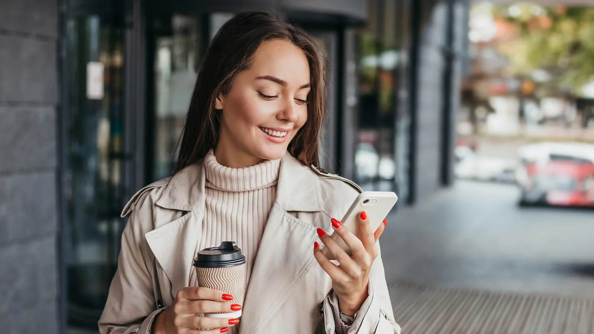 Young smiling ukrainian brunette woman holding a smartphone and a cup of coffee looking at device screen against the background of an office building. copy space