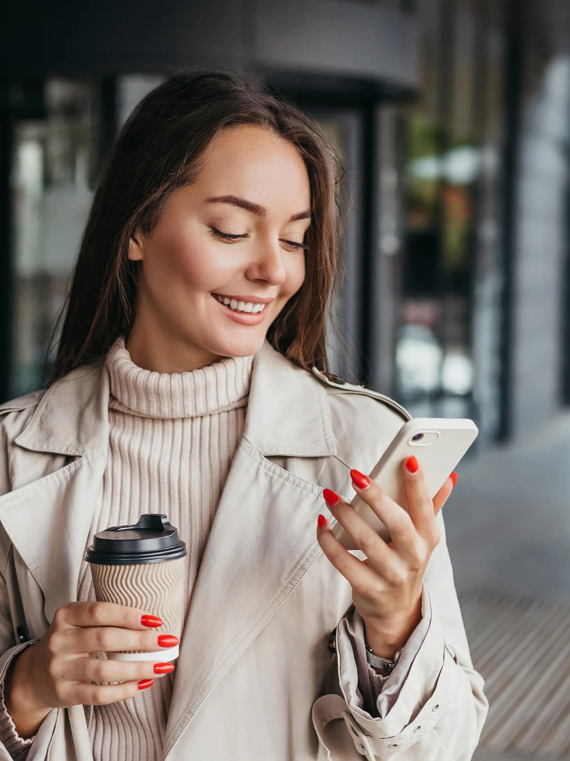 Young smiling ukrainian brunette woman holding a smartphone and a cup of coffee looking at device screen against the background of an office building. copy space