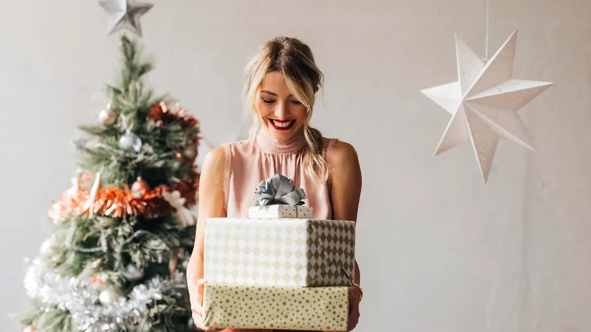 Cheerful smiling female standing next to the decorated Christmas tree and holding wrapped gift boxes in hands
