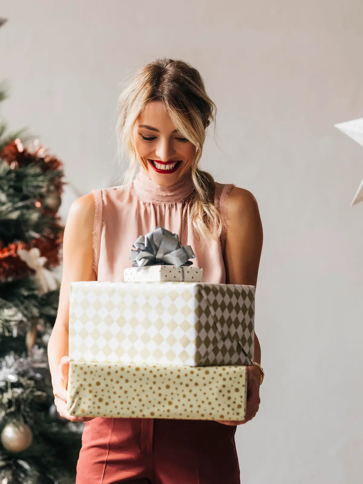 Cheerful smiling female standing next to the decorated Christmas tree and holding wrapped gift boxes in hands