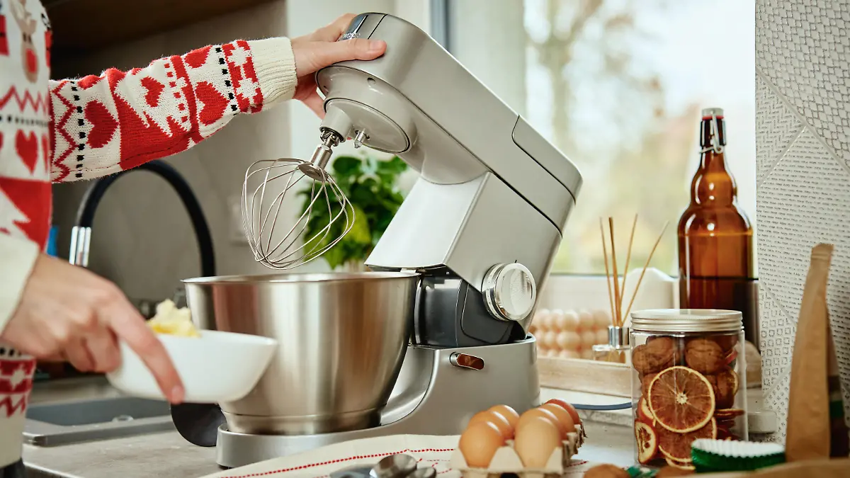 Woman in festive christmas sweater preparing dough for cookies at home kitchen. Female hands use electric mixer. Modern kitchen household appliances