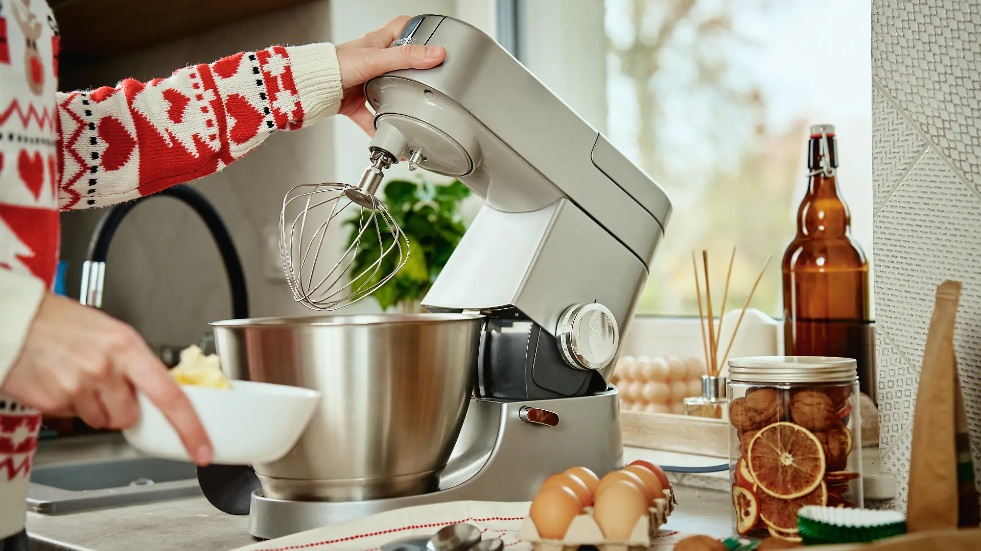 Woman in festive christmas sweater preparing dough for cookies at home kitchen. Female hands use electric mixer. Modern kitchen household appliances