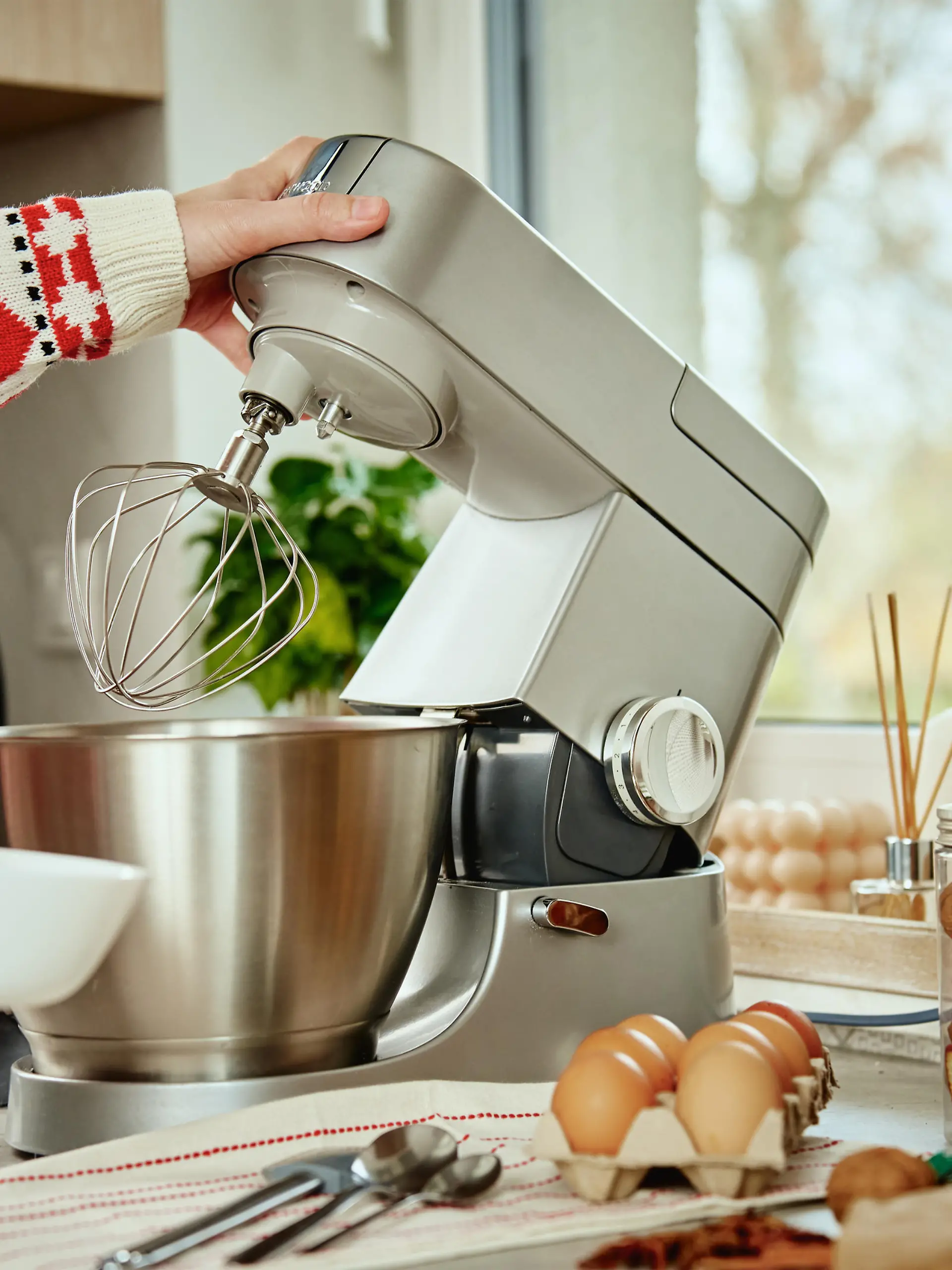 Woman in festive christmas sweater preparing dough for cookies at home kitchen. Female hands use electric mixer. Modern kitchen household appliances