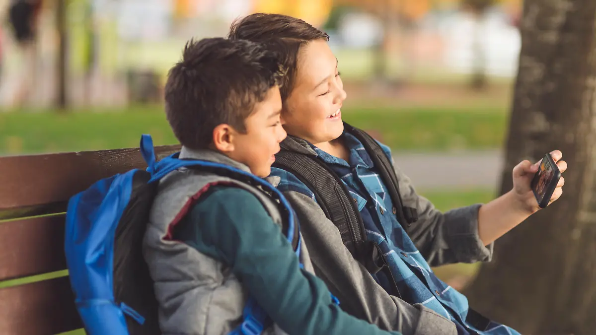 Two elementary-age brothers sit on a park bench in the late afternoon and watch a funny video on their phone. They both have backpacks and are smiling.