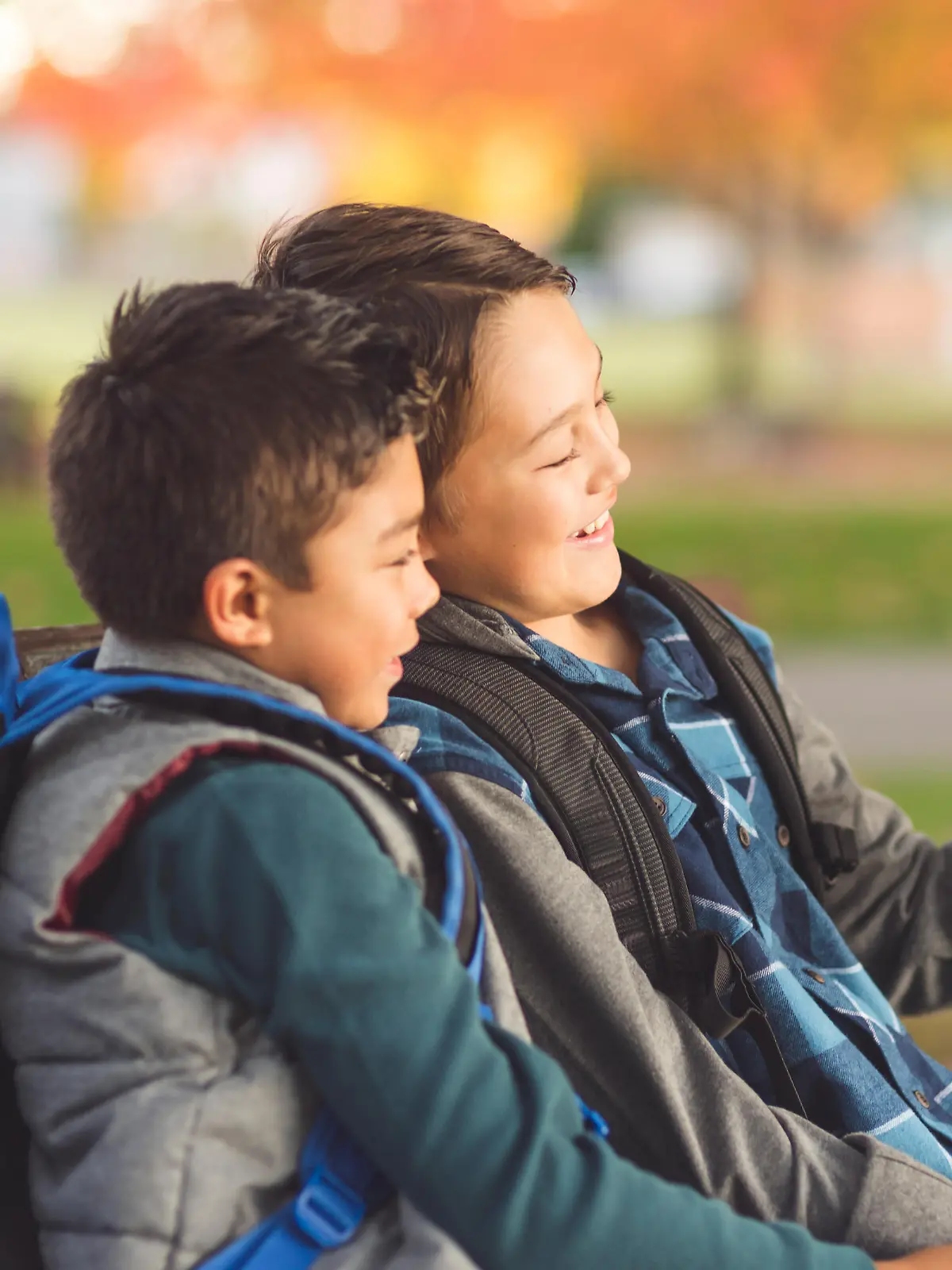 Two elementary-age brothers sit on a park bench in the late afternoon and watch a funny video on their phone. They both have backpacks and are smiling.