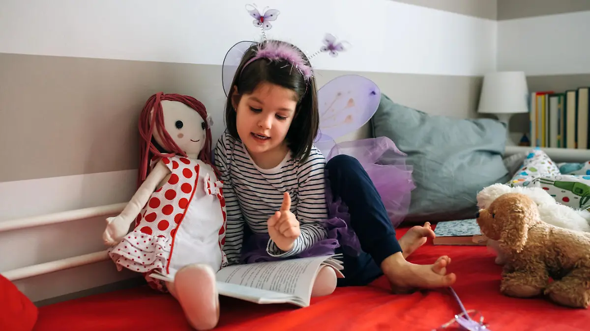 Little girl disguised as a butterfly sitting on the bed reading a book to her rag doll