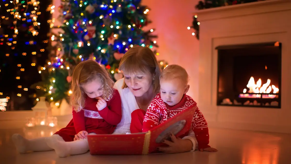 Mother and children read a book at fireplace on Christmas eve. Family with child celebrating Xmas. Decorated living room with tree, fire place and candles. Winter evening at home for parents and kids.