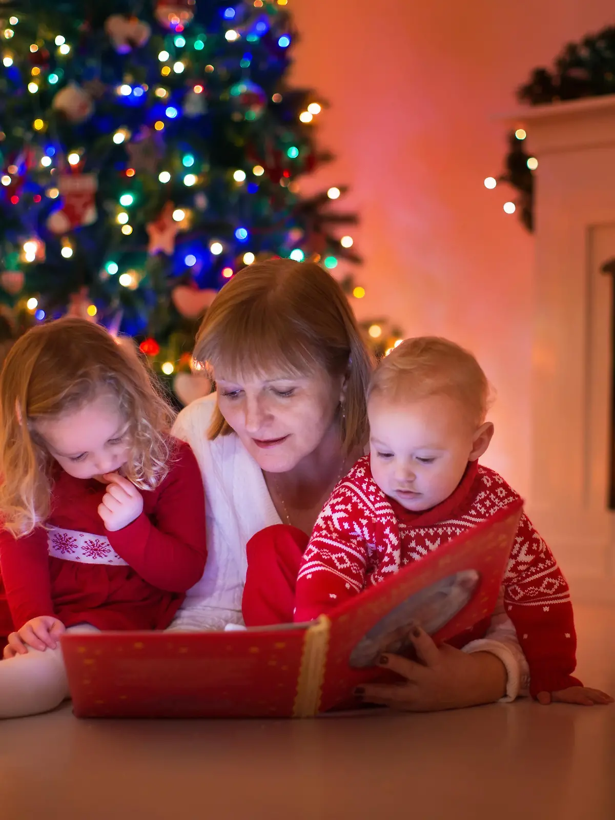 Mother and children read a book at fireplace on Christmas eve. Family with child celebrating Xmas. Decorated living room with tree, fire place and candles. Winter evening at home for parents and kids.