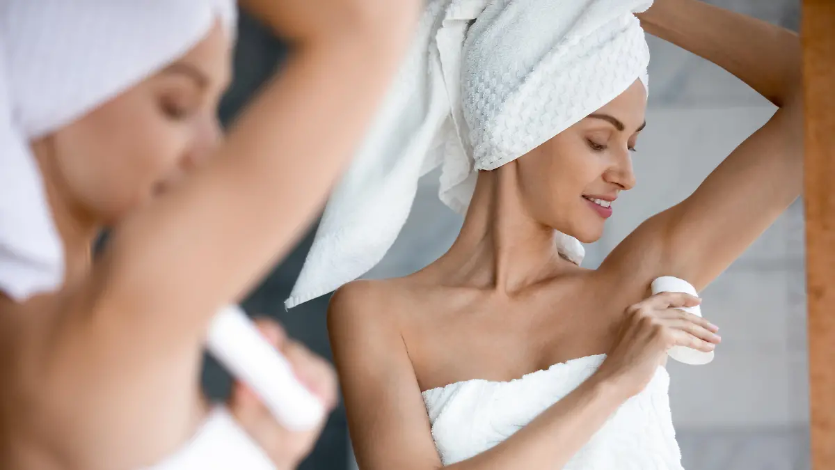 Smiling beautiful woman applying antiperspirant to armpit after shower close up, standing in front of mirror in bathroom, pretty young female wearing white bath towel on head using stick deodorant