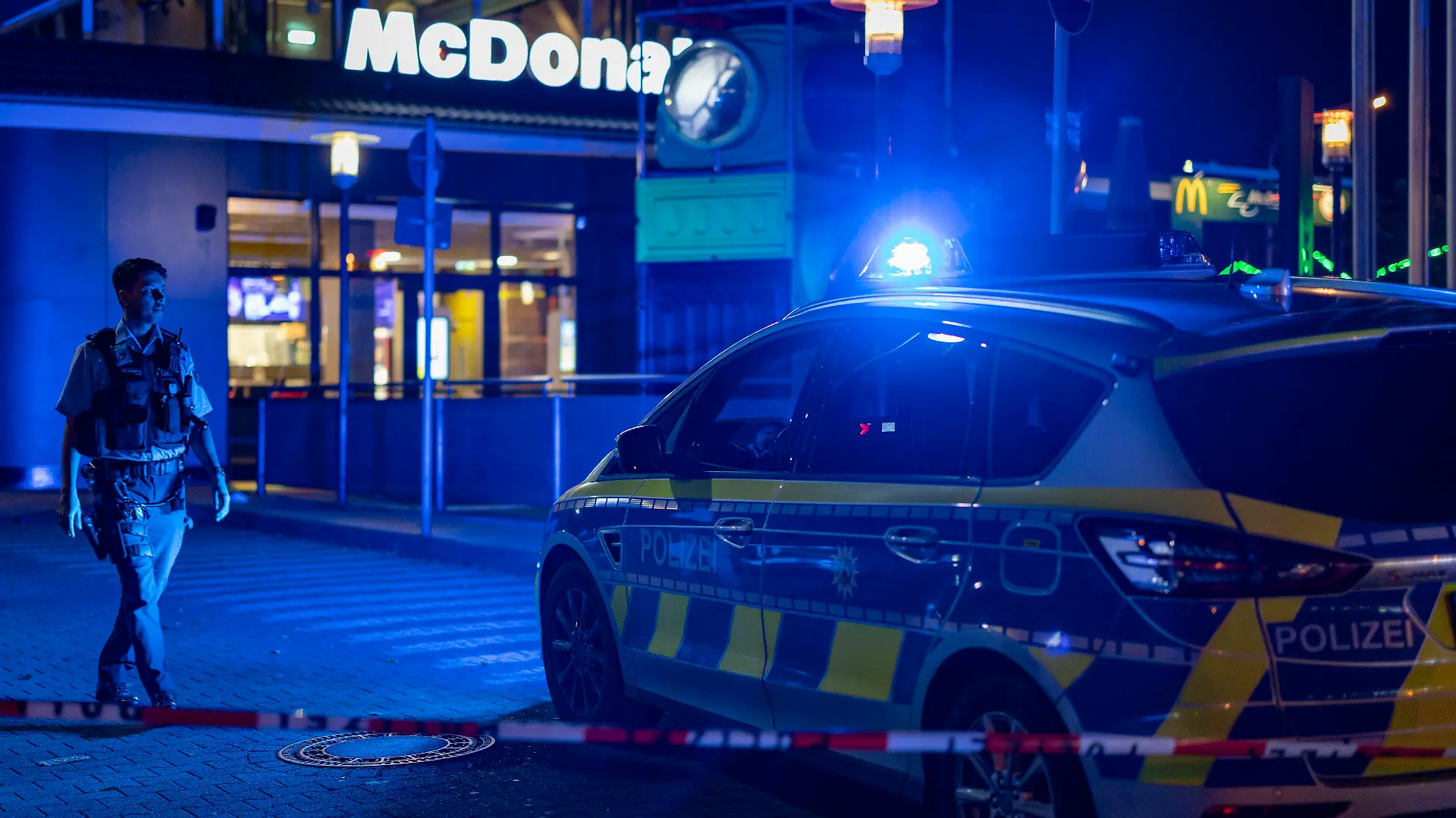 29.10.2022, Nordrhein-Westfalen, Oberhausen: Ein Polizist geht vor einem Fast-Food-Restaurant im Blaulicht, nachdem im Außenbereich Schüsse gefallen waren. Drei Menschen wurden verletzt. Der Täter sei auf der Flucht, teilte ein Polizeisprecher mit. Die Hintergründe der Tat sind dem Sprecher zufolge noch unklar. Foto: Christoph Reichwein/dpa +++ dpa-Bildfunk +++