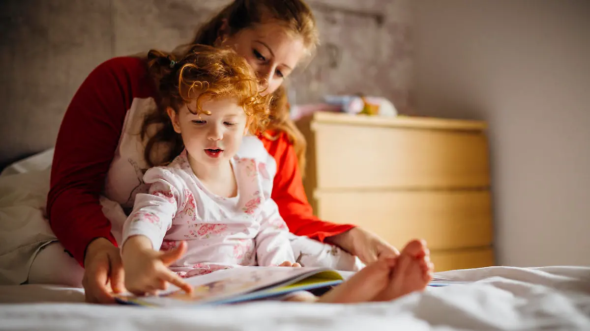 Little girl reading a bedtime story with her Mother in her bed. They are sitting together and the little girl is pointing at one of the pictures.